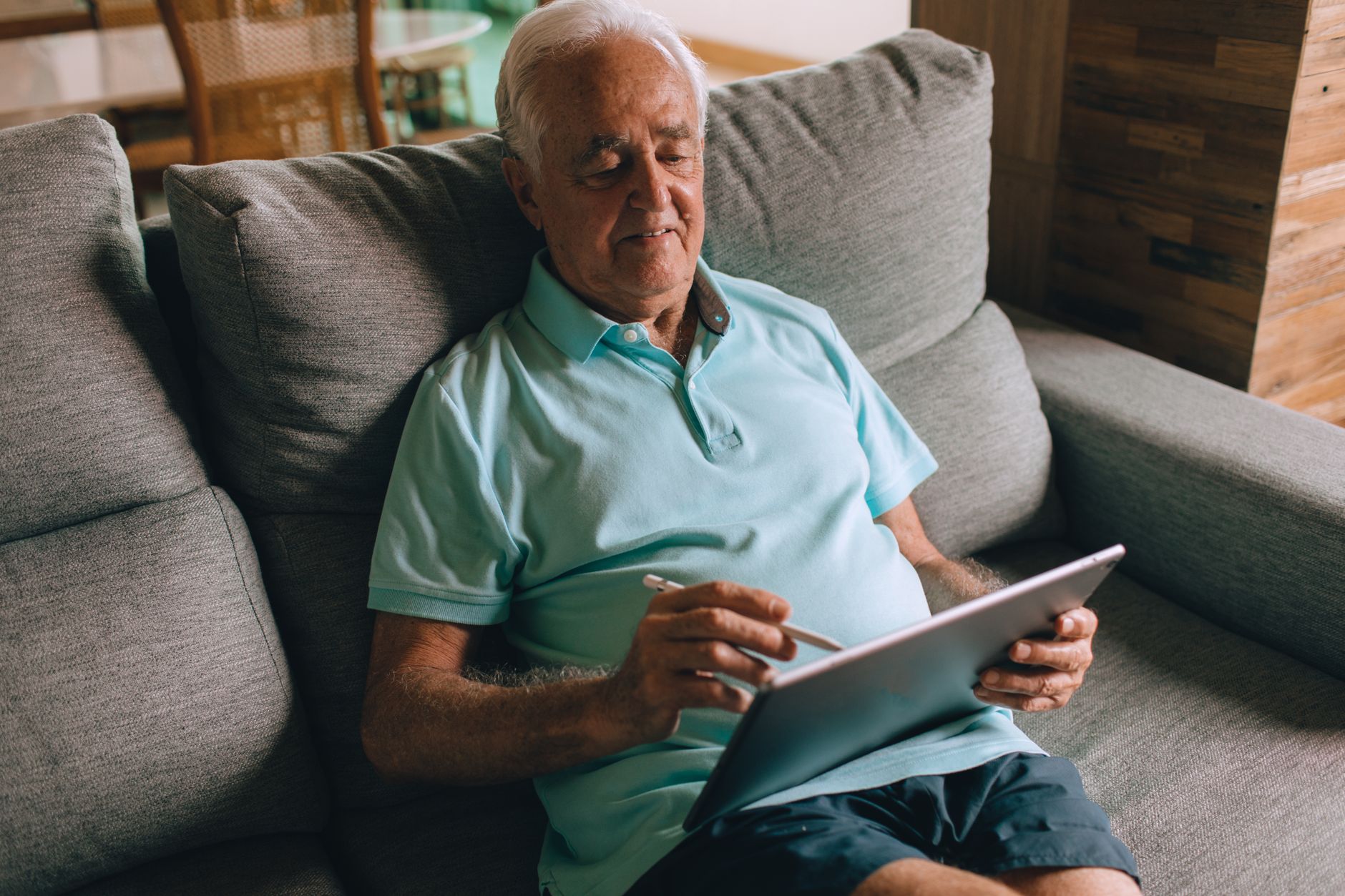Homme senior en polo bleu assis sur un canapé, utilisant une tablette avec un stylet à l'intérieur.