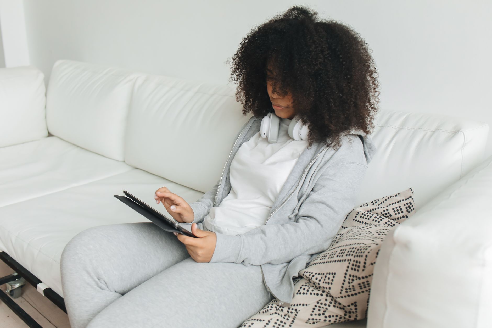 Femme avec un casque utilisant une tablette sur un canapé blanc, tenue décontractée grise.